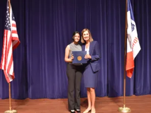 A young woman with Governor Kim Reynolds, standing between an American flag and a State of Iowa flag, holding a Governor's Volunteer Award folder between them.