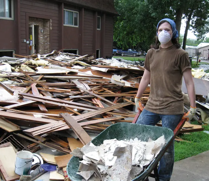 Person wearing a facemask holding a wheelbarrow in front of a pile of debris
