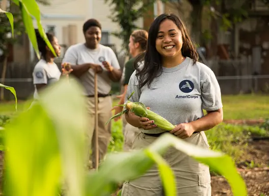 Young woman wearing grey t-shirt with AmeriCorps logo on it holding an ear of corn