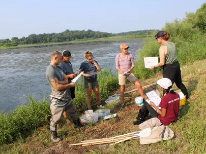 A group of young adults standing along a riverbank, organizing a clean up